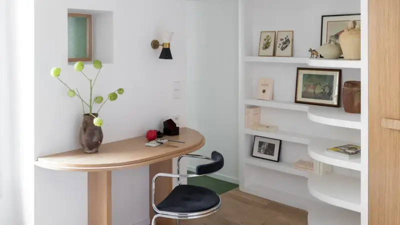 Un despacho pequeño en casa con un escritorio curvo de madera, una silla de terciopelo azul y estanterías blancas llenas de libros y objetos decorativos.